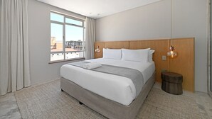 A hotel room with a large bed, a wooden headboard, a small wooden side table, a window with curtains, and a city view. - Elegant One Bed Sea Point Mountain Views (Cape Town)