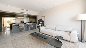 A modern living room with a sofa, coffee table, and bar stools. A kitchen is visible in the background. - Elegant One Bed Sea Point Mountain Views (Cape Town)