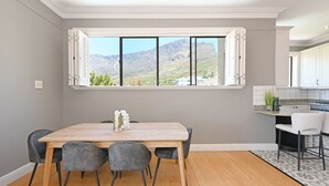 A dining area with a wooden table and grey upholstered chairs, a window with a mountain view, and a kitchen with white cabinets. - Two Bed Table Mountain Spacious (Cape Town)