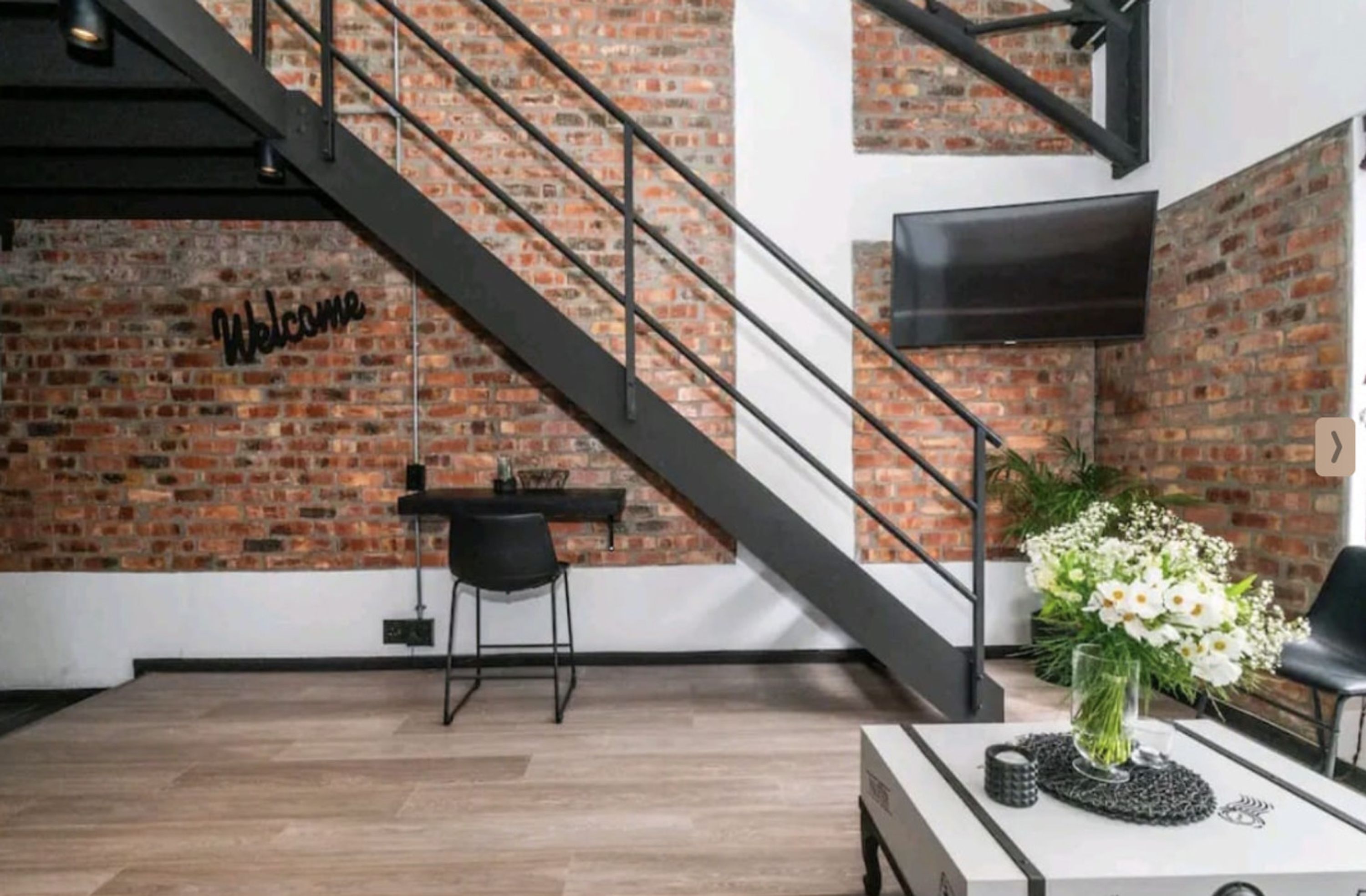 A modern interior with a brick wall, a staircase, a flat-screen TV, and a table with a vase of flowers.