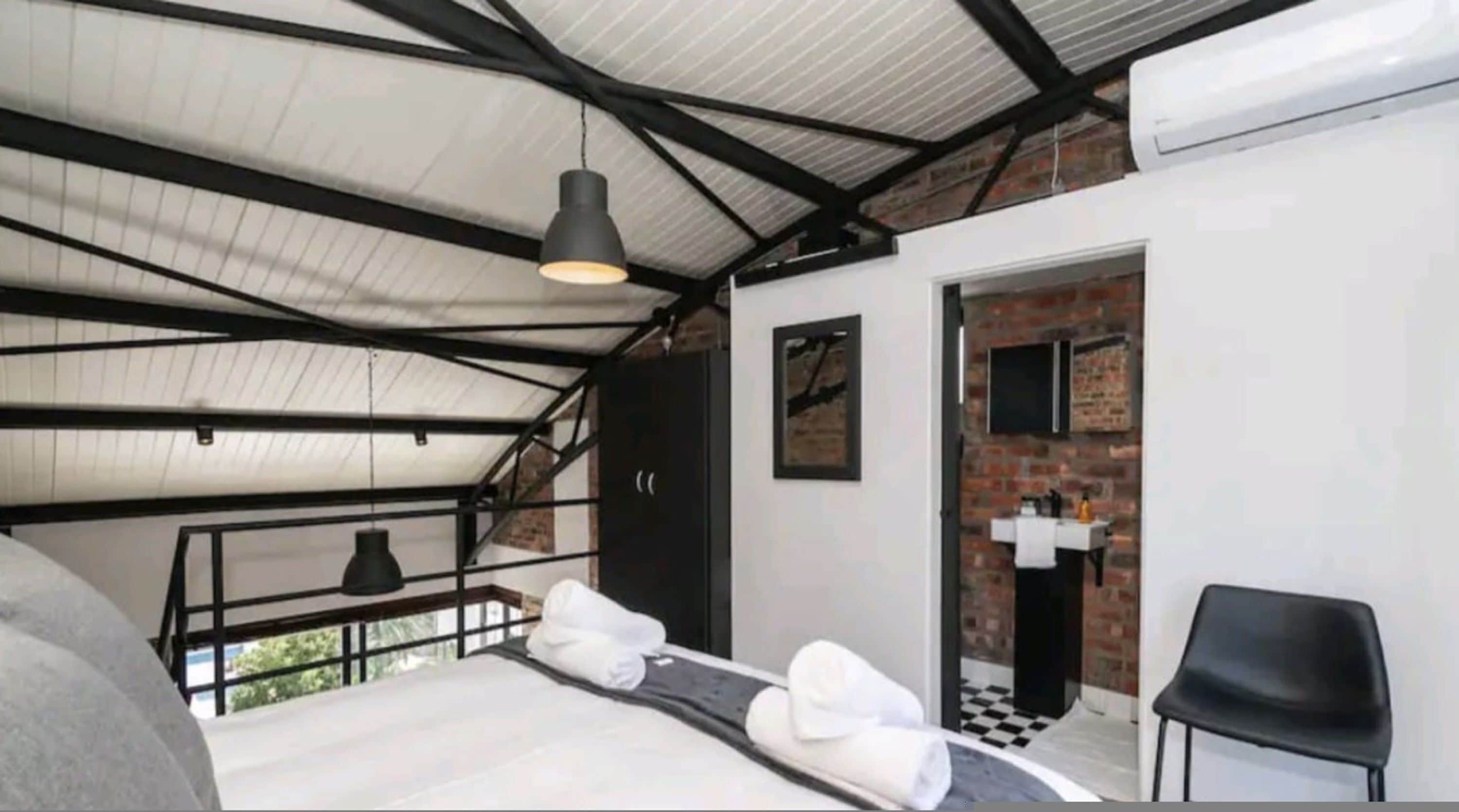 A bedroom with a bed, a black chair, a hanging light fixture, and a door leading to a bathroom with a sink and mirror.