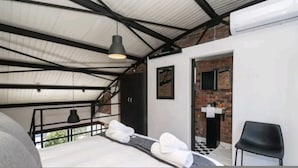 Quarto com cama, cadeira preta, luminária pendente e porta para banheiro com pia e espelho.