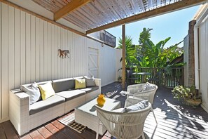 Une terrasse extérieure couverte, aménagée avec un canapé en osier, des fauteuils en osier blancs et une petite table sur laquelle est posé un vase jaune.