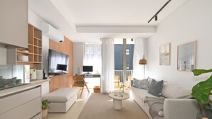 En moderne stue med sofa, sofabord og fjernsyn. Der er et køkkenområde med skabe og et spisebord.