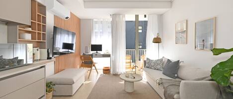 A modern living room with a sofa, coffee table, and TV. There is a kitchen area with cabinets and a dining table.