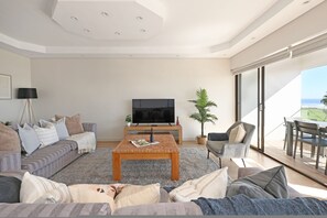 Una sala de estar moderna con televisor de pantalla plana, una mesa de centro, sofás y un comedor con vista al océano.