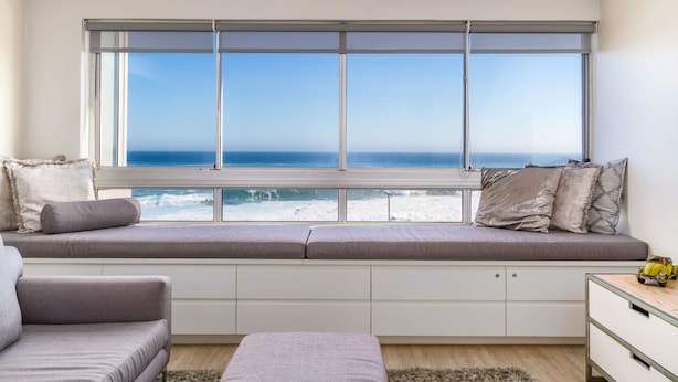 Una habitación moderna con un ventanal grande con vistas al océano, una banca empotrada con cojines y una pequeña mesa con un juguete.