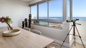Una habitación moderna con un amplio ventanal con vista al océano, un telescopio sobre un trípode, una mesa de madera con un plato decorativo y un jarrón con flores.