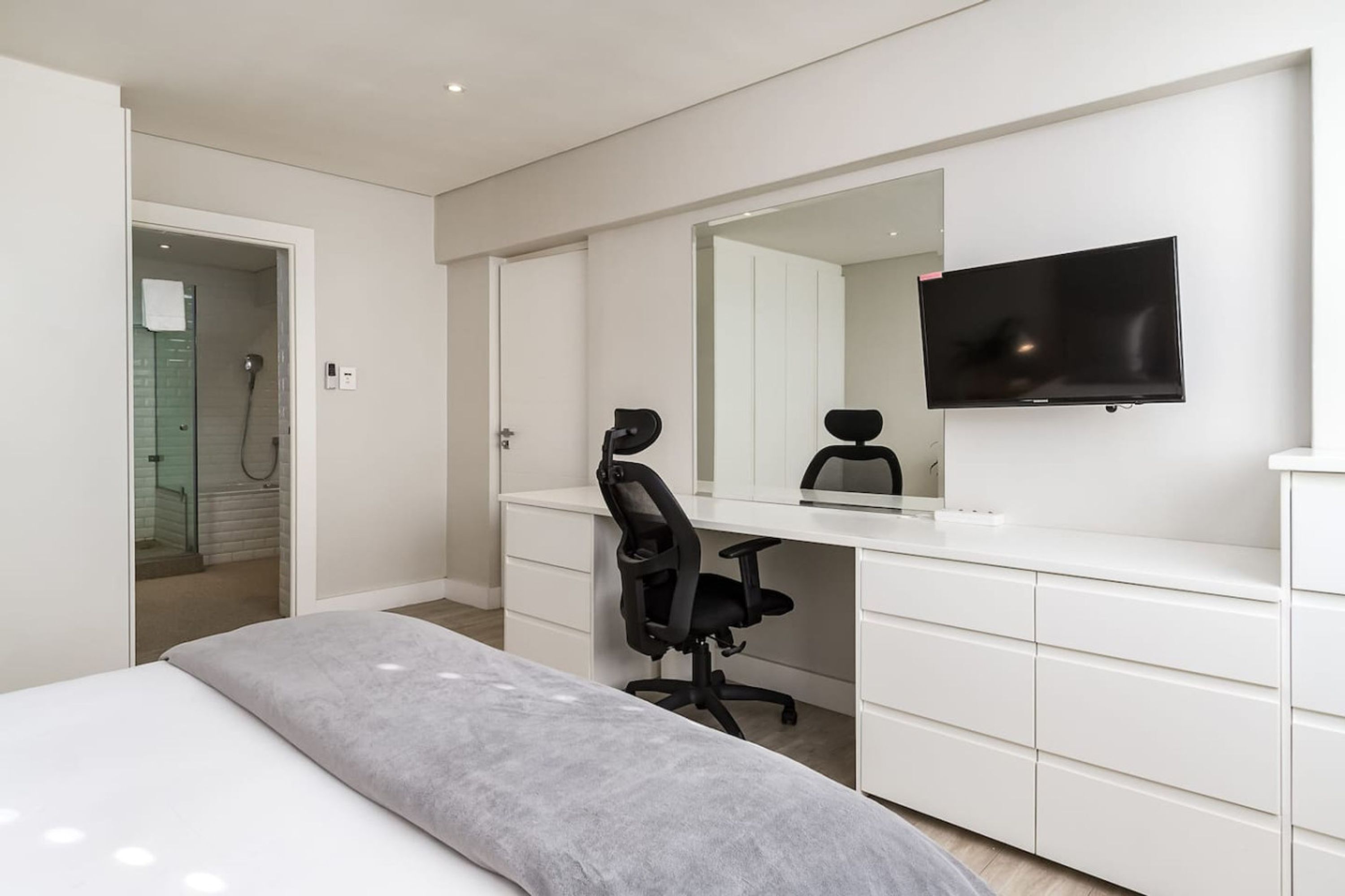 Et moderne hotelværelse med seng, skrivebord med stol, stort spejl og fladskærms-tv.