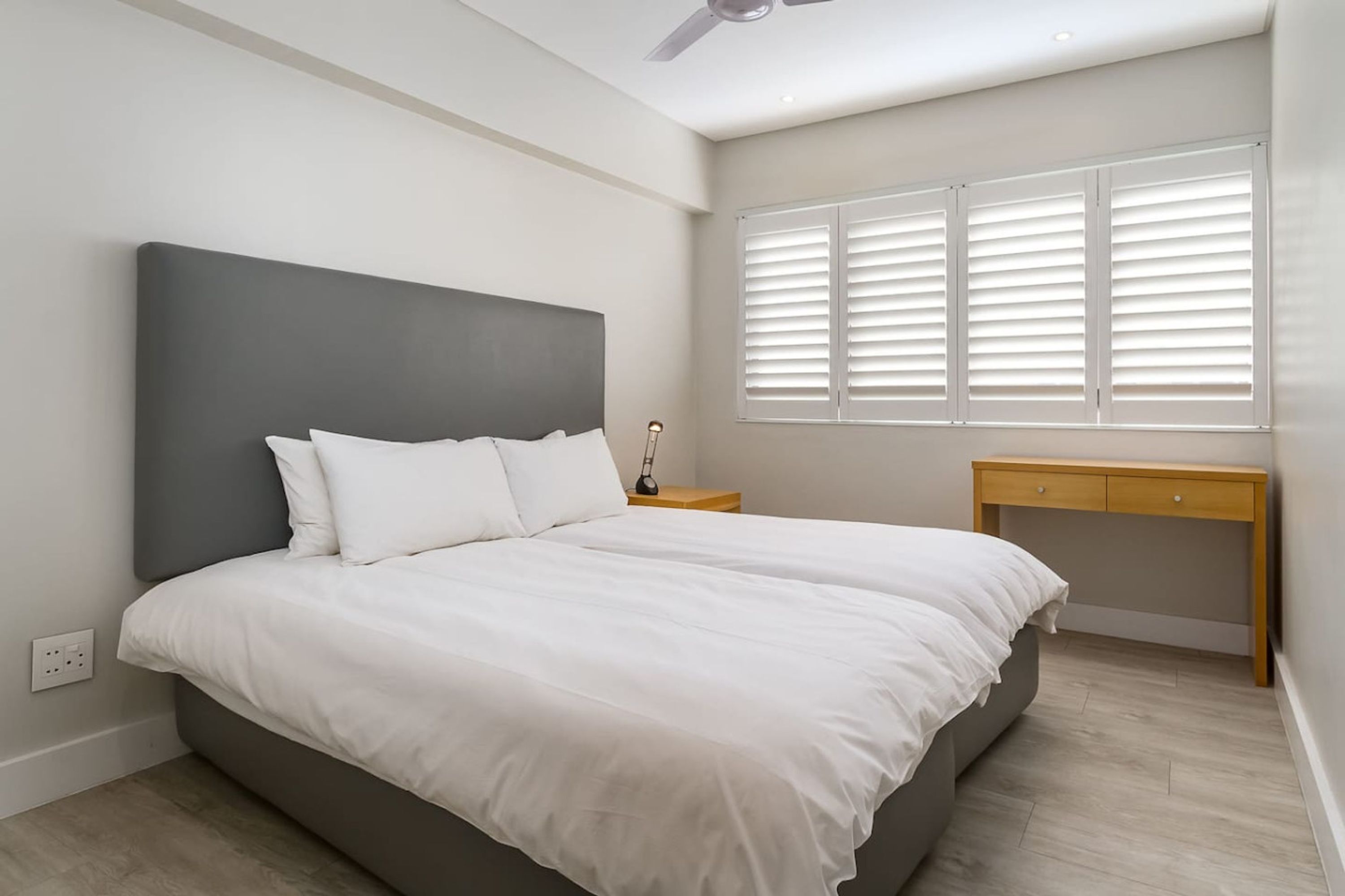 A bedroom with a large bed, a bedside table, a desk, and a window with blinds.