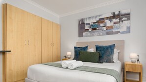 Een slaapkamer met een grote houten garderobe, een bed met witte lakens, twee nachtkastjes met lampen en een modern abstract schilderij aan de muur.