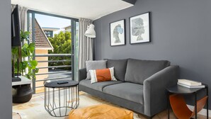 A modern living room with a grey sofa, a small round table, a single orange chair, and a large window with a view of buildings and trees. - Planet Africa Studio Pool Gym (Cape Town)