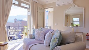 Une pièce avec un canapé beige, une petite table et une chaise. Il y a un balcon d’où l’on peut admirer les bâtiments et les montagnes.