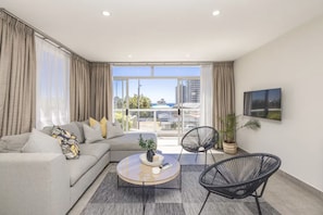 A modern living room with a sofa, coffee table, and chairs, large windows with curtains, and a view of buildings and greenery. - Seaview Bliss a Sunny Apartment and Pool (Cape Town)