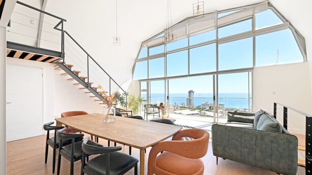 A spacious living area with a large window overlooking the sea, a wooden dining table with chairs, and a modern sofa. - Spacious 3 Bedroom Villa BBQ Pool Ocean View (Cape Town)