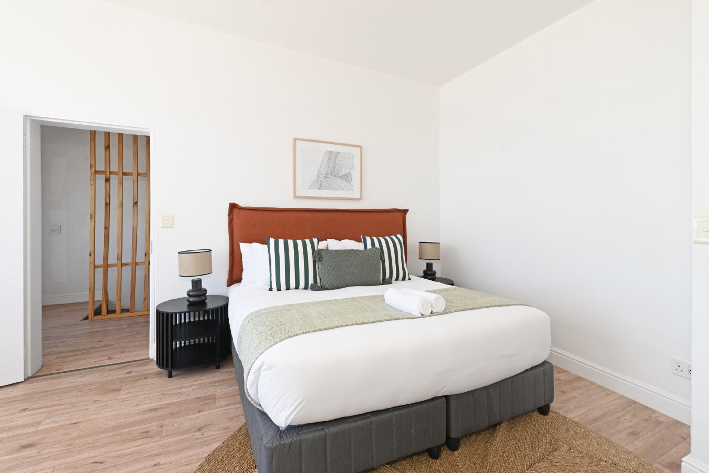 Un dormitorio con cama, mesitas de noche, una puerta y un cuadro colgado en la pared.