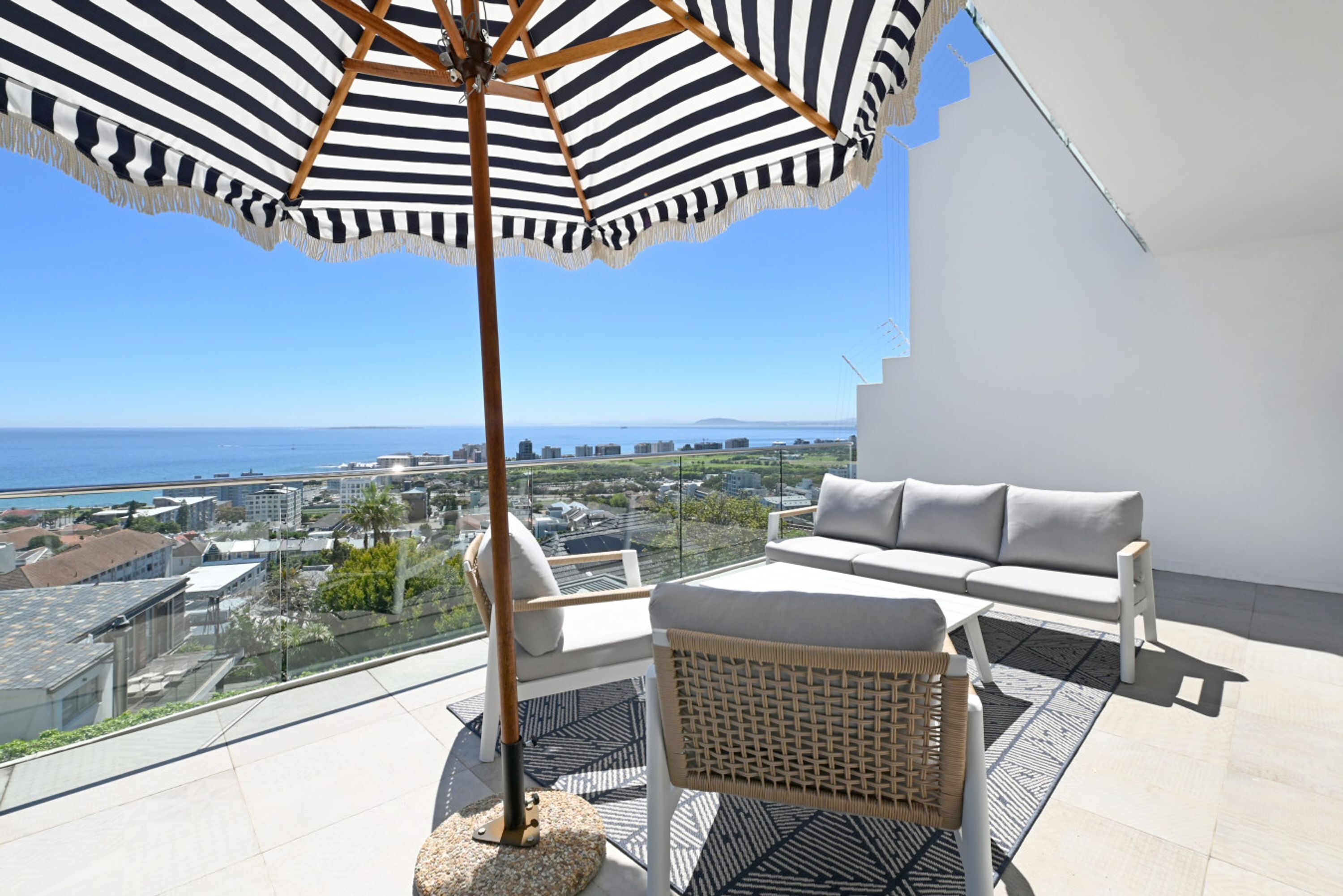 A balcony with a view of the sea, a striped umbrella, a sofa, and a wicker chair.