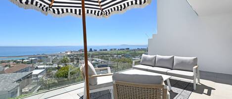A balcony with a view of the sea, a striped umbrella, a sofa, and a wicker chair.