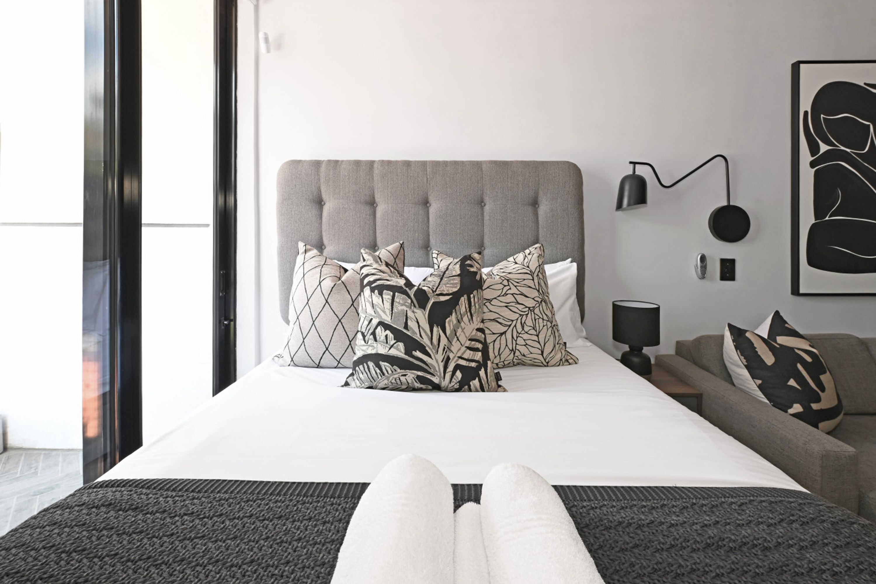 A modern hotel room with a large bed, a grey upholstered headboard, and a grey sofa.