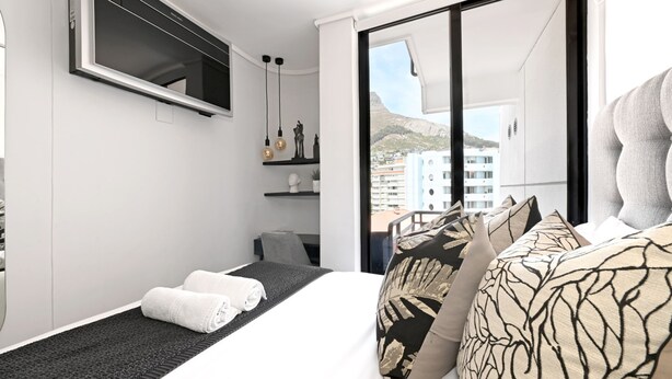 Una habitación de hotel moderna con una cama grande, un televisor montado en la pared, un balcón con vistas a edificios y una pequeña zona de estar con una silla y una mesa.
