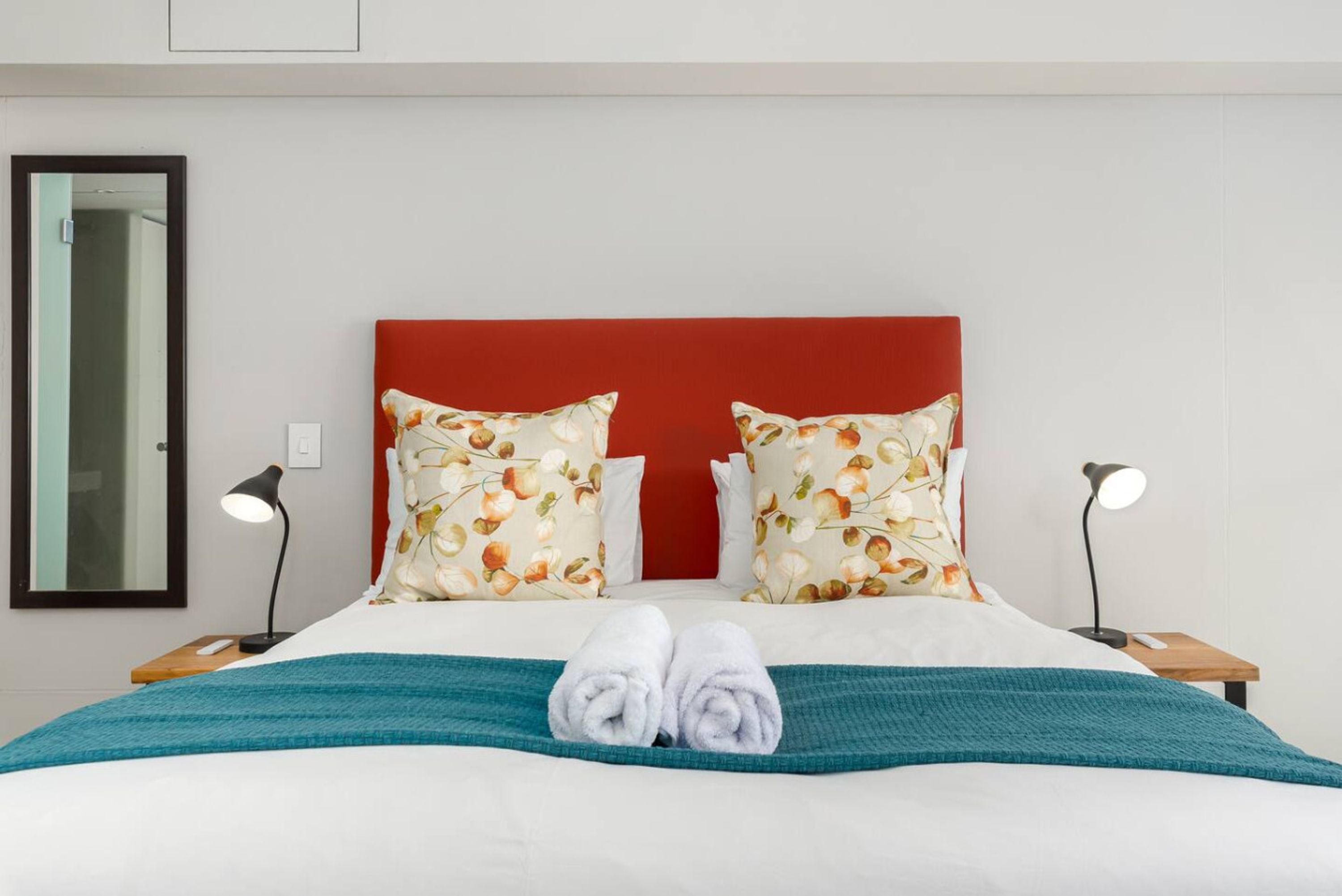 A neatly made bed with a red headboard, floral pillows, and a teal blanket. There are two bedside tables with lamps.