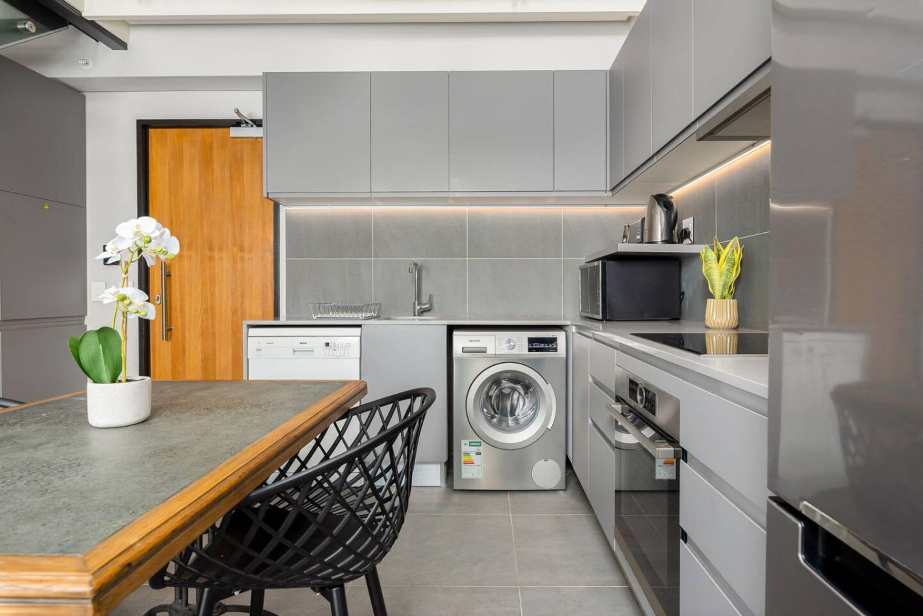 Una cocina moderna con armarios grises, lavadora integrada, microondas y una mesa de madera con una silla.