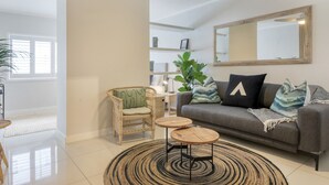 Un salon moderne avec un canapé gris, un fauteuil en osier, deux petites tables rondes en bois, un grand miroir et une plante en pot.