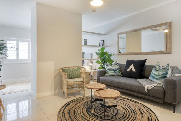 A modern living room with a grey sofa, a wicker chair, two round wooden tables, a large mirror, and a potted plant. - Chic Sea Point Retreat Walk to the Promenade Caf s (Cape Town)