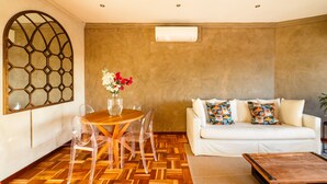 A modern living room with a wooden herringbone floor, a round wooden table with chairs, a white sofa with colorful cushions, and a large decorative window. - Modern One Bed Central Green Point Stadium (Cape Town)
