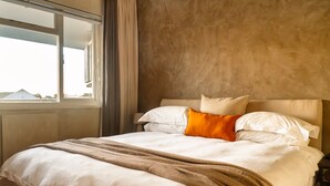 A neatly made bed with white linens and a brown throw blanket, a headboard, and a window with curtains. - Modern One Bed Central Green Point Stadium (Cape Town)