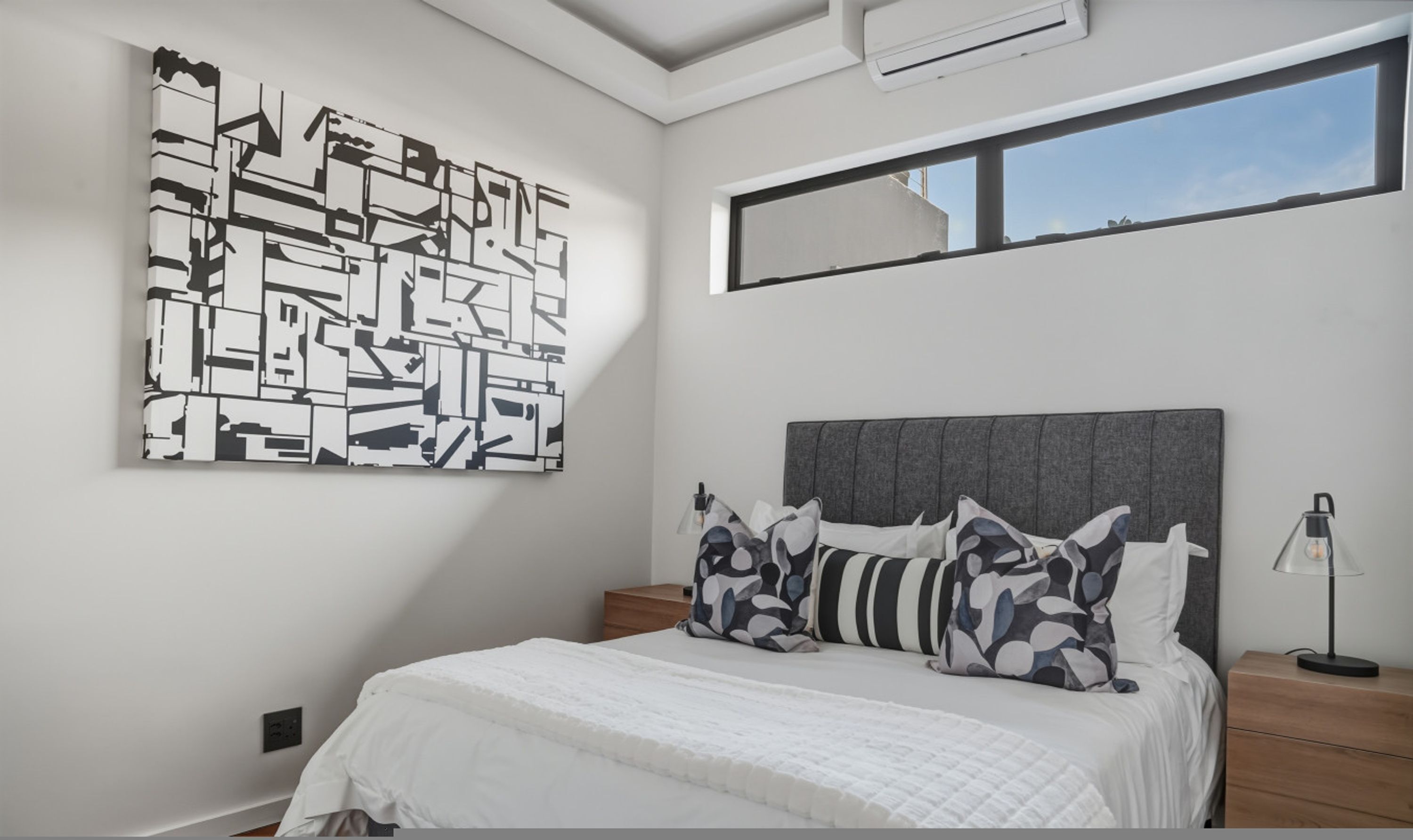 A bedroom with a large abstract painting, a grey headboard, and a bedside table with a lamp.