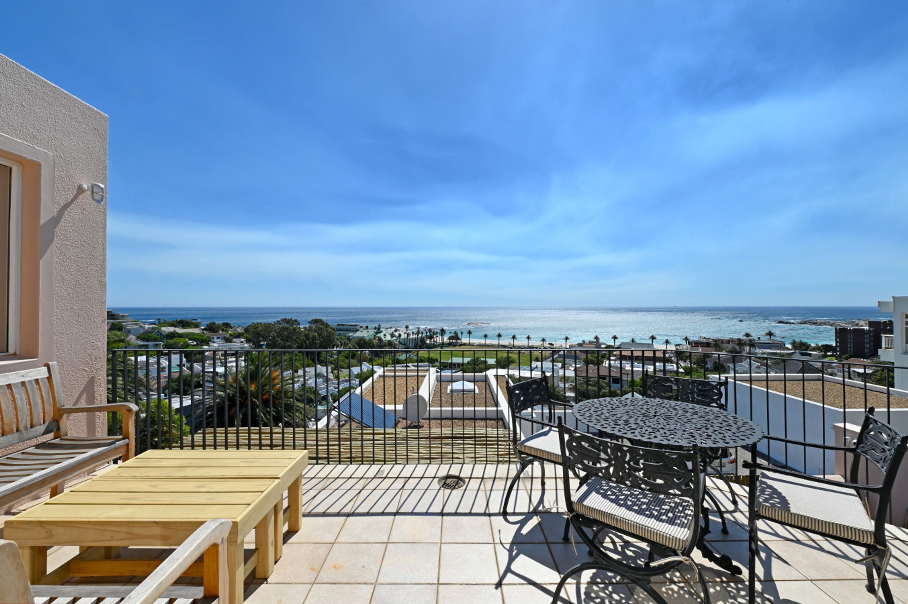 A balcony with a wooden bench, a round table with chairs, and a view of the ocean and cityscape.