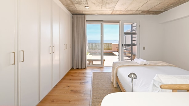 A bedroom with a wooden floor, a bed with white linens, a large window with a view of the sea, and a sliding glass door leading to an outdoor area. - Elegant Camps Bay Eco Pool Sauna Views (Cape Town)