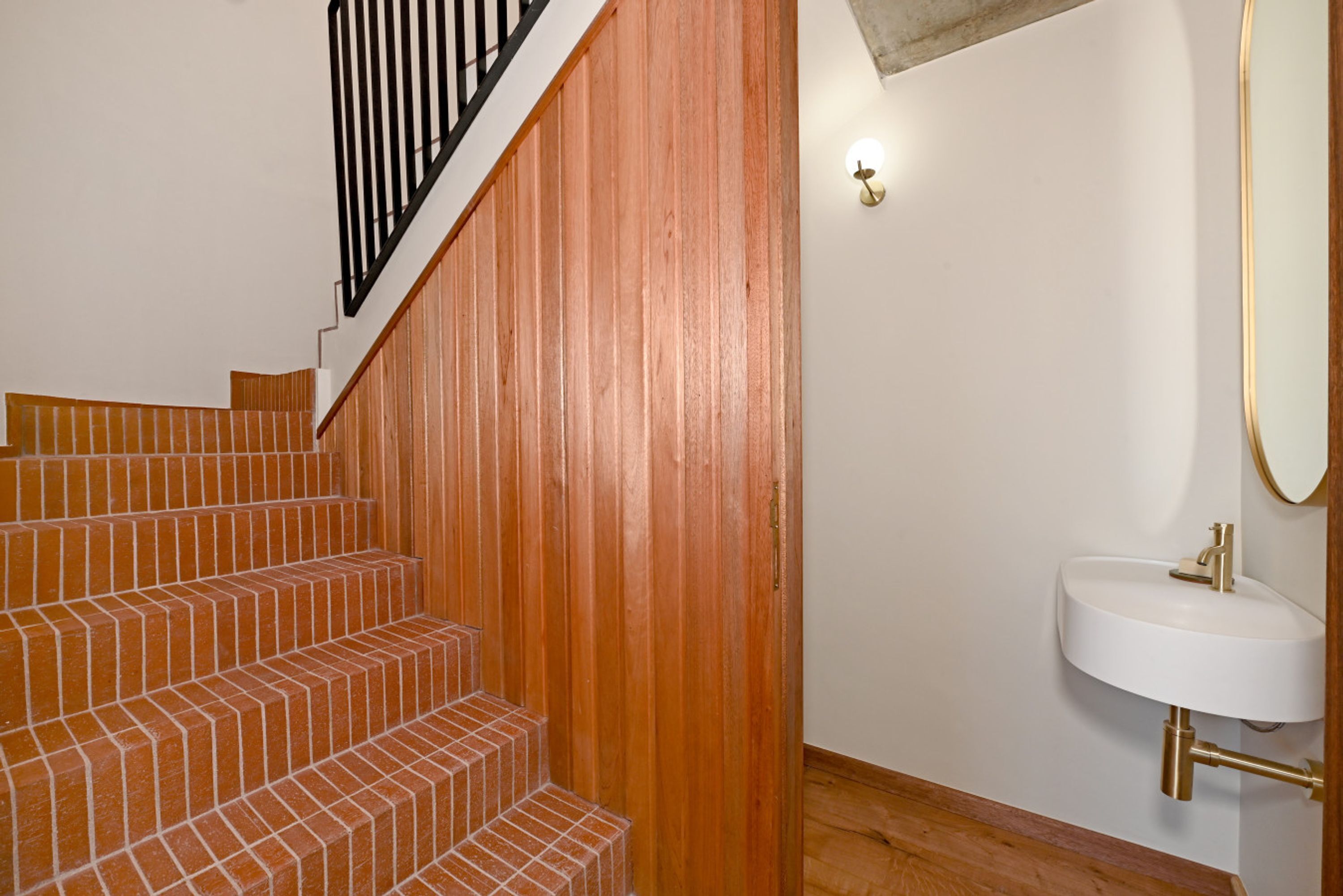 A staircase with wooden steps and a black railing, leading to a white wall with a gold-colored sink and a mirror.