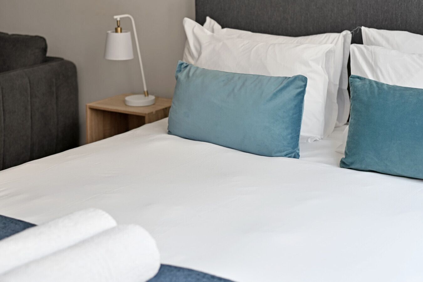 A neatly made bed with white and teal pillows, a wooden nightstand with a white lamp, and a grey upholstered armchair.