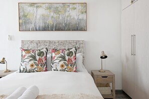 A bedroom with a bed, two pillows with floral patterns, a bedside lamp, a wall-mounted painting, and a wardrobe. - Secure Sea Point Spacious Three Bedroom House (Cape Town)