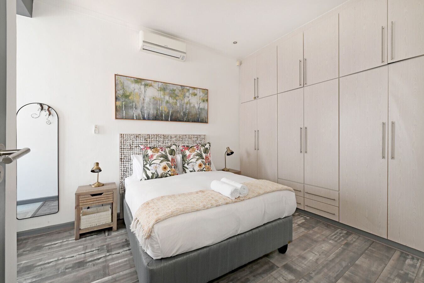 A modern bedroom with a bed, a nightstand, a mirror, a wardrobe, and an air conditioning unit.