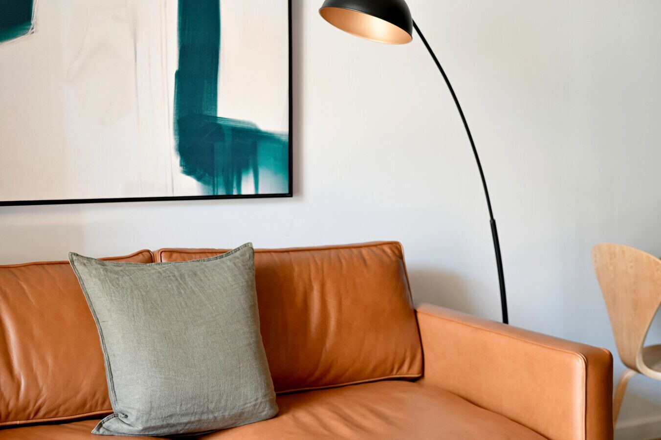 A modern living room with a leather sofa, a grey pillow, a black floor lamp, and a wooden chair.
