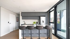 Egy modern konyha szürke szekrényekkel, egy központi szigettel, amelyhez bárszékek tartoznak, és egy nagy ablakkal, melyből kilátás nyílik a külvilágra.