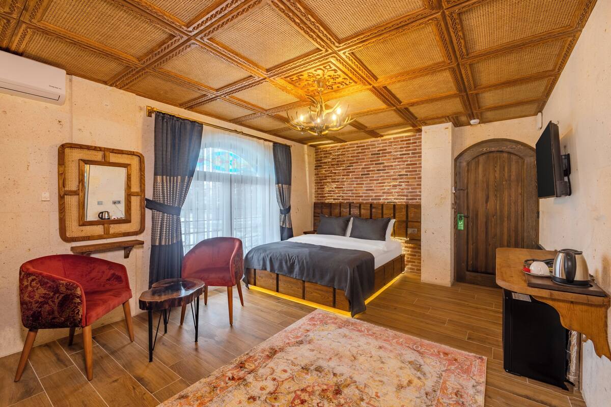 A bedroom with a wooden ceiling, a brick wall, a bed, a red chair, a small table, and a television on the wall.