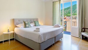 Een slaapkamer met een bed, nachtkastje, stoel en een balkon met uitzicht op gebouwen en heuvels.