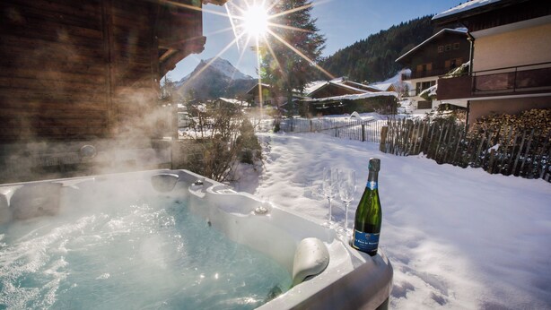 Uma banheira de hidromassagem com uma garrafa de vinho e copos em um deck coberto de neve, rodeado por edifícios de madeira e montanhas.