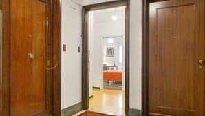 Un couloir avec des portes en bois, un paillasson et une pièce visible où l’on aperçoit un couvre-lit orange.
