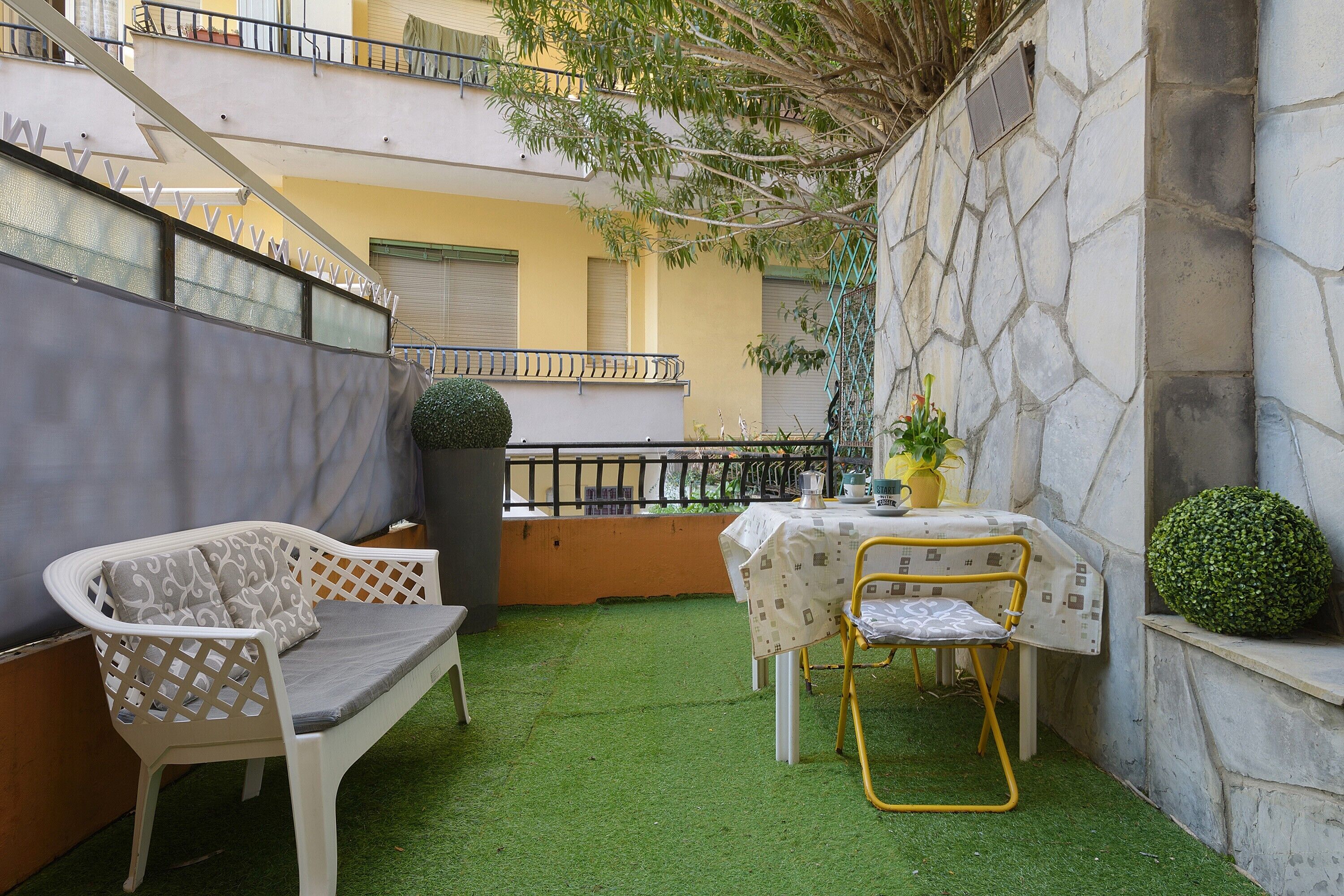 En lille udendørs terrasse med en hvid sofa, en gul stol, et lille bord og en potteplante.