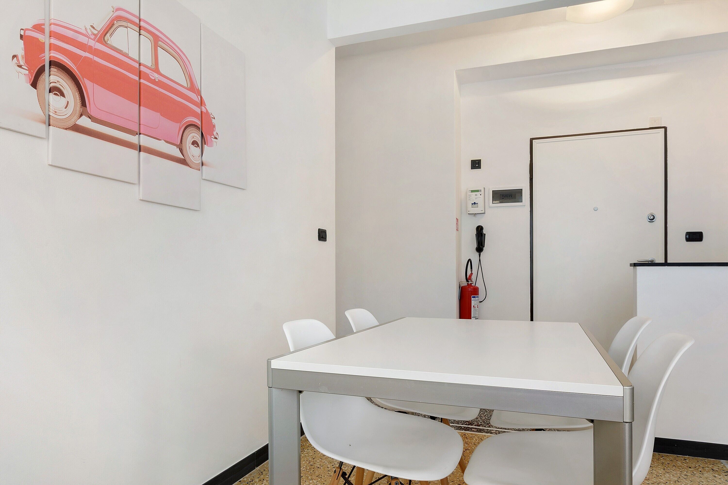 A conference room with a white table and chairs, a fire extinguisher, and a door in the background.