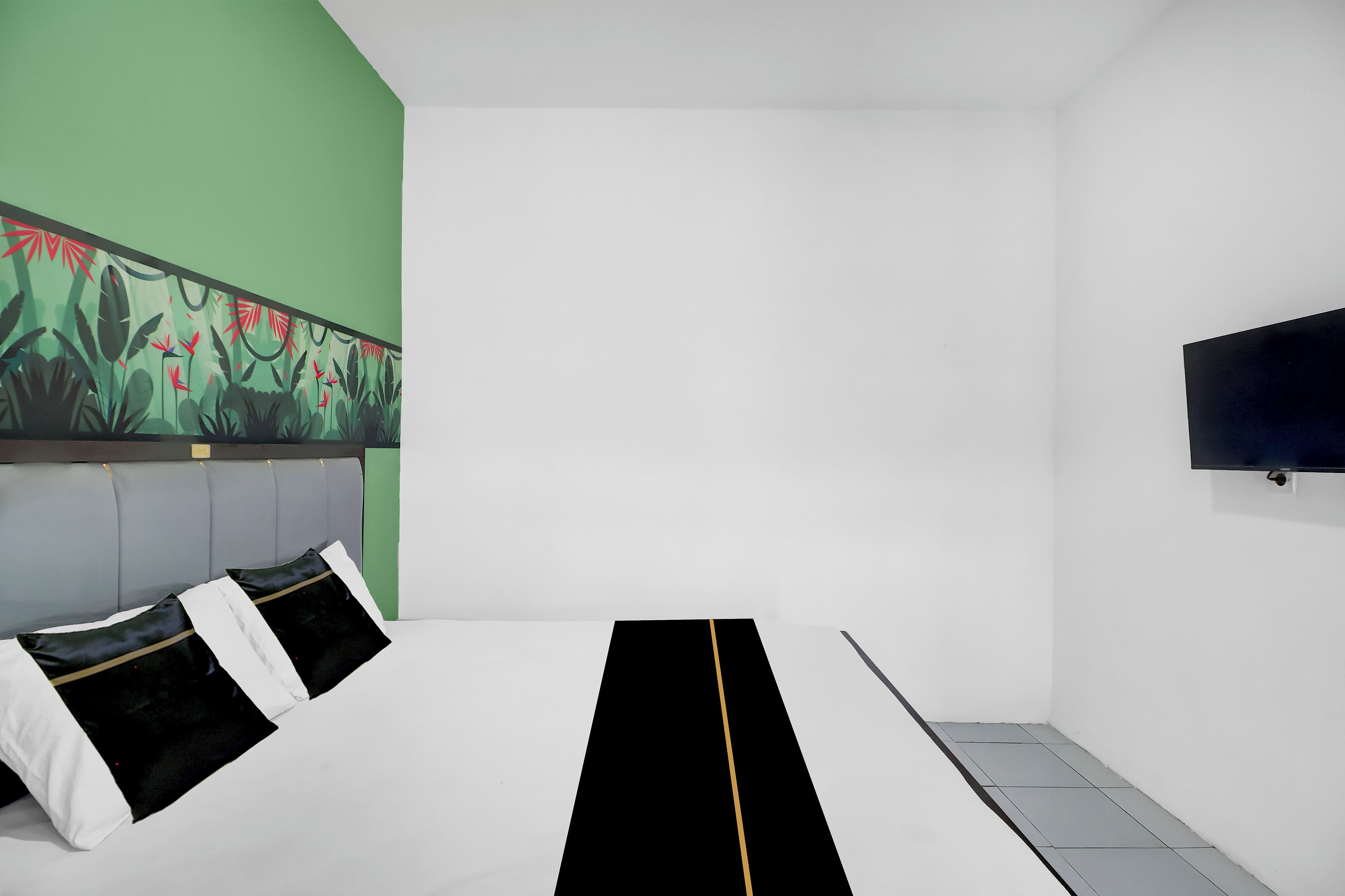 A hotel room with a bed, a green wall with a floral mural, a flat-screen TV mounted on the wall, and a tiled floor.
