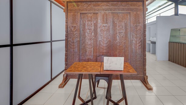 A room with a traditional wooden partition featuring intricate carvings, a wooden table with a cushion, and a modern ceiling with a hanging light fixture. - Hotel O near Trans Studio Theme Park formerly Sunset Cozy (Kuta)