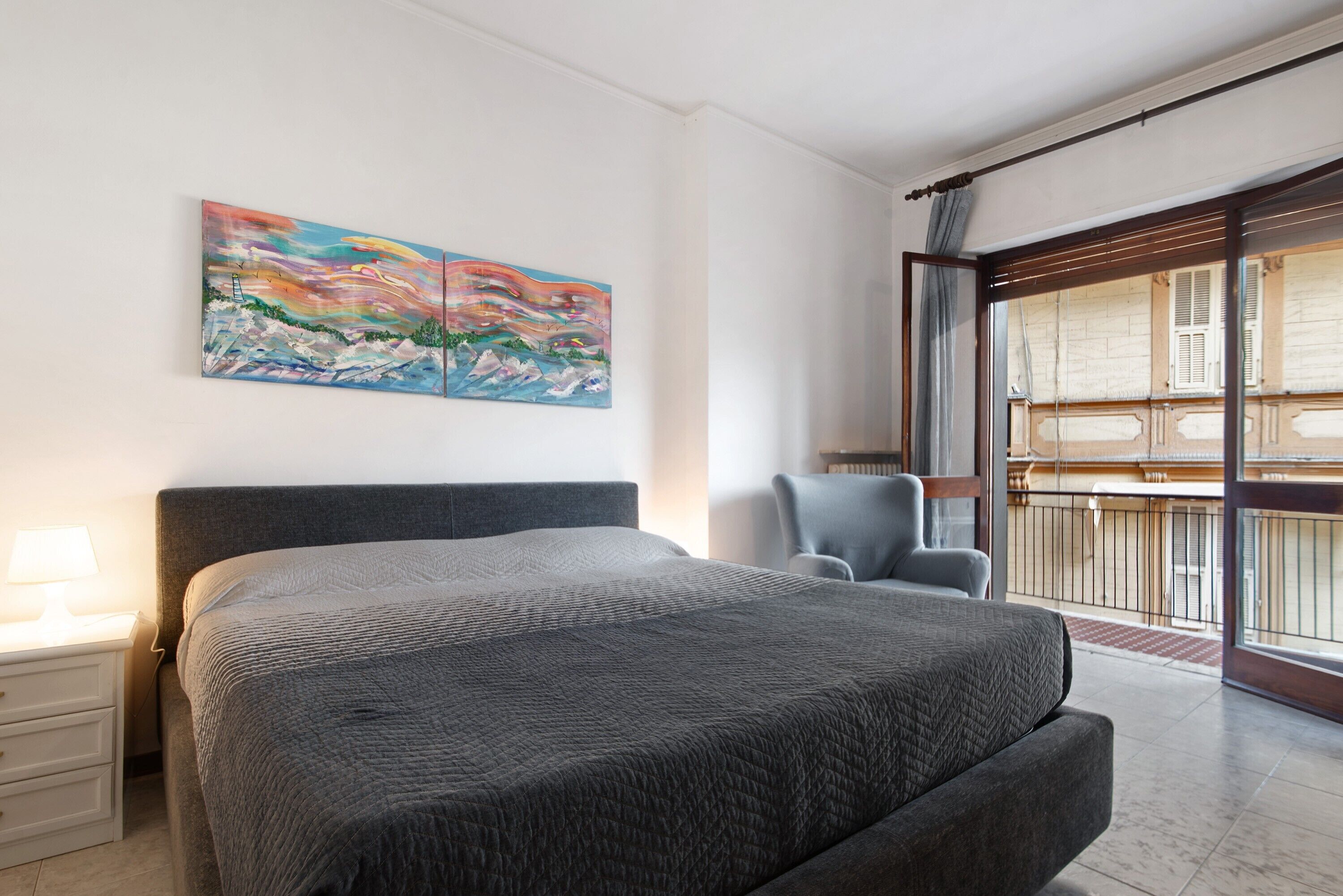 Un dormitorio con cama, una silla, una mesita de noche, un cuadro y un balcón.