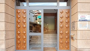 Une entrée d’immeuble avec une porte en verre, une porte en bois ornée de motifs en losange et un escalier menant à l’intérieur.