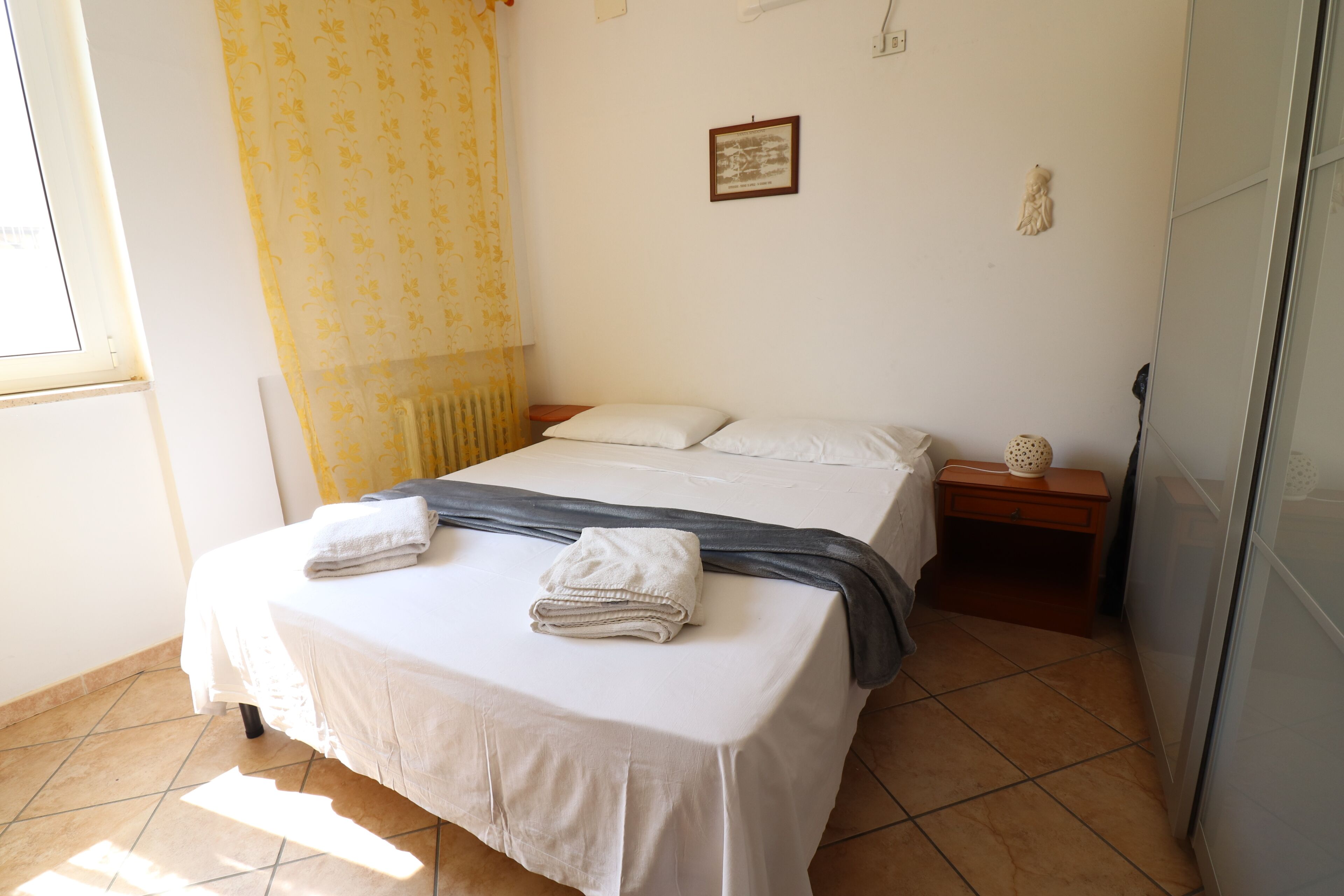 A neatly made bed with white linens and a gray blanket, a small bedside table with a lamp, and a window with yellow curtains.